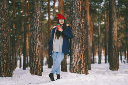 Caucasian girl in red hat with paper cup stands near the tree looks into the distance. Walking in...