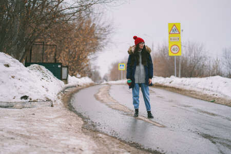 Depressing view of a muddy dirty road with melting snow and road signs. Girl in red hat stands in...