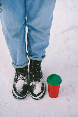 Red paper cup with green lid stands on the snow near human feet. Do not litter concept. Recycle