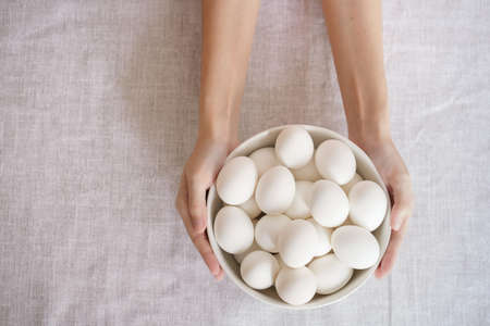 Thin female hands holding white bowl of eggs on the table. Easter or healthy food conceptの写真素材