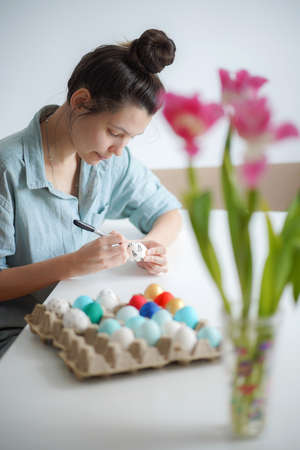 Caucasian girl with hair bun is preparing for Easter drawing on eggs. Bokeh pinik flowers. Colorful...