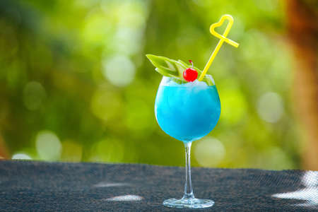 Horizontal picture of blue cocktail with apple, cherry and yellow straw in wine glass on background of green bokeh. Bartender skills conceptの写真素材