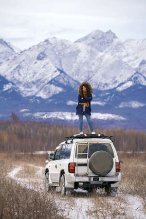 Car trip to snowy mountains. Girl with dreadlocks stands on white off road car roof with her arms crossed. Siberian or European landscapeの写真素材
