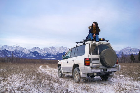 Car trip to snowy mountains. Girl with dreadlocks sits on white off road car roof. Siberian or European landscapeの写真素材