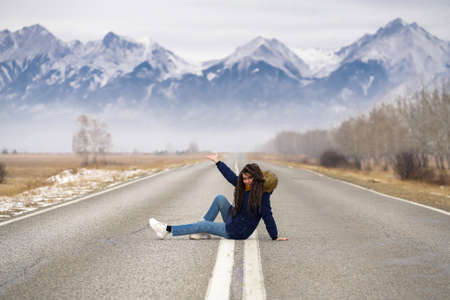 Travel to mountains. Caucasian girl with dreadlocks sits in the middle of road showing mountains.の写真素材