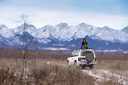 Car trip to snowy mountains. Girl with dreadlocks sits on white off road car roof and holds her...