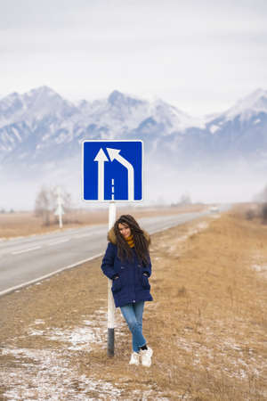 Travel destination. Caucasian girl with dreadlocks leans on blue road sign on snowy mountains...