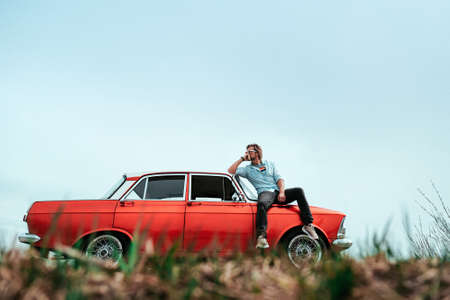 Young man in sunglasses sitting on the hood of his red retro car. Concept of free life