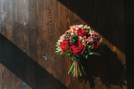 Colorful wedding bouquet on table made of dark wood in rays of bright sun. Red and pink roses with small tulips. The play of light and shadow.の写真素材