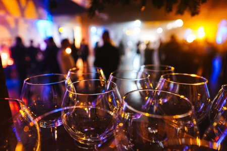 Wine glasses in the light of multicolored lights against the background of blurred silhouettes of people. Large-scale party or private event with catering servicesの写真素材