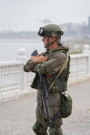 Kaspiysk, Dagestan, Russia - July 25, 2021: Young man in a military uniform on embankment with an arms around a machine gun rifle looks somewhere in the distance with mouth open.のeditorial素材
