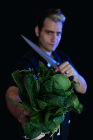 Vegan chef holds a bundle of raw spinach and a cleaver in his other hand with a ring, looking defiantlyの写真素材
