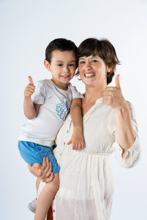 Young mother and son standing together on white background making happy thumbs up hand gesture. approving expression looking at camera showing success.の写真素材