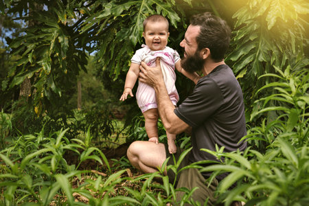 A happy family. Dad and baby play and laugh on a walk. A baby girl looks at the camera smiling, her father holds her and they are surrounded by green leafy plants. They have a great time together.の写真素材