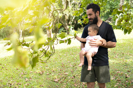 Dad and baby in the park walks through a park among green bushes and trees, a baby girl looks and touches the green leaves of the trees.の写真素材