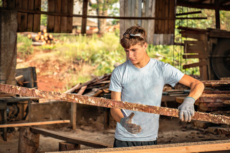Young man working in a sawmill wearing protective gloves. Young teenage boy carrying boards of wood freshly cut by a machine.の写真素材