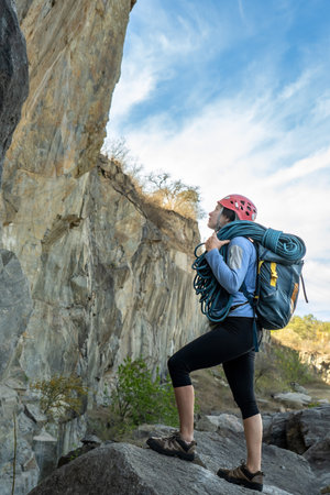 Female climber standing in front of a rock, carrying climbing rope, looking up, observing rocky mountain.の写真素材