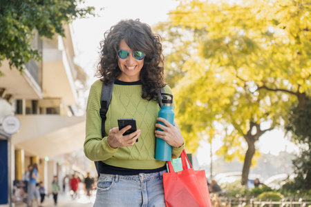 Smiling woman with curly hair walks through the city at sunset, holding a water bottle and a red...