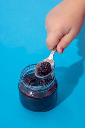 Kids hand holding spoon with berries. Jar of Blackberry Jam on blue paper. Selective focus.の写真素材