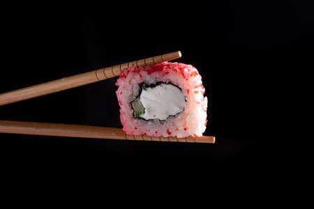 Chopsticks holding seaweed pink rice roll filled with avocado and cheese over dark background, copy spaceの写真素材