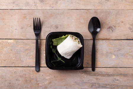 tasty fresh wrap sandwich with chicken and vegetables in black container on wood table, spoon and fork near. Flat layの写真素材