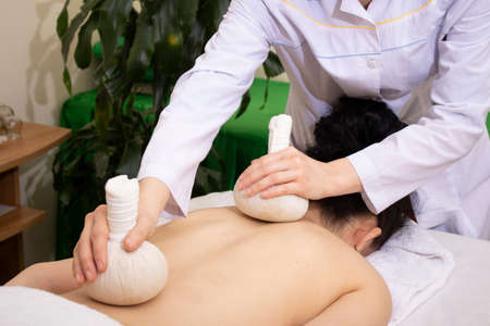 Herbs tea scrub in spa salon, massager holding two pouchesの写真素材