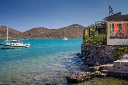 Cafe on the shore at the water's edge. Two boats are moored side by side. Low mountains in the background. Gulf of Mirabello. Elounda. Crete. Greece.の写真素材