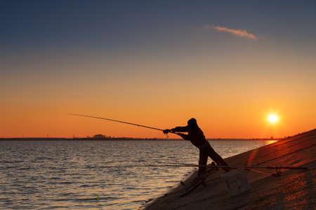 Silhouette of fisherman fishes on river bank against a sunsetの写真素材