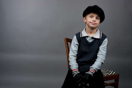 young caucasian old fashioned boy sitting on chair wearing gloved and cap isolated over gray backgroundの写真素材