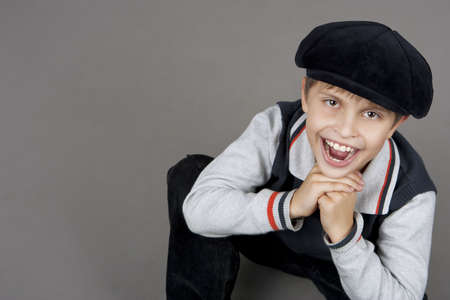 young cute and smiling happy teenager boy sitting on floor with old cap and looking up having fun with mouth open and hands support chin isolated over gray backgroundの写真素材