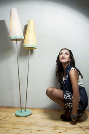 happy bright brunette with excited facial expression sitting near high lamp in front of gray wallの写真素材