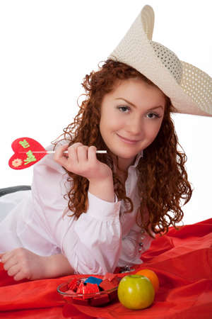 young beautiful caucasian red haired girl wearing big hat and collecting candies lying on floor upon red linen and having positive natural smile isolatedの写真素材