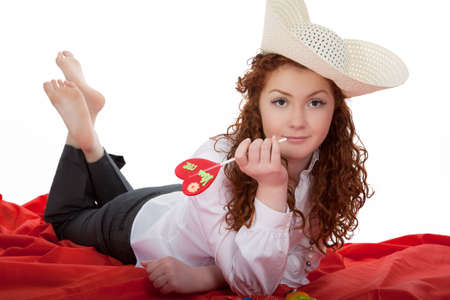 relaxing and dreaming young red haired caucasian girl wearing big hat and holding heartilke bon-bon in hand lying on red linen isolated on whiteの写真素材
