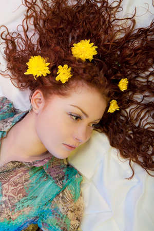extravagant and bright looking caucasian red haired beautiful girl having yellow flowers interlaced in her hair lying over white riffled linen with calm and dreaming facial expressionの写真素材