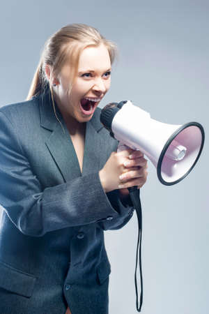 Furious Caucasian Blond Female in Suite Shouting Using Megaphone. Against Gray Background. Vertical Imageの写真素材