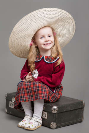Happy And Smiling Caucasian Child Posing in Big Round Sombrero Hat and Sitting on Outdated Suitcase. Eating Chocolate Sweet. Against Gray Background. Vertical Imageの写真素材
