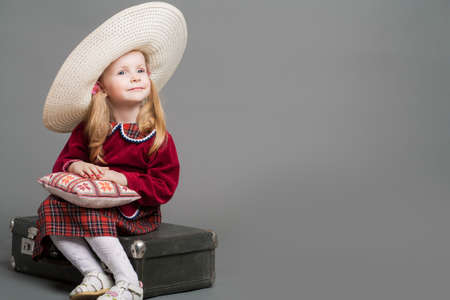 Happy And Smiling Caucasian Child Posing in Big Round Sombrero Hat and Sitting on Outdated Suitcase. Against Gray Background. Horizontal Imageの写真素材