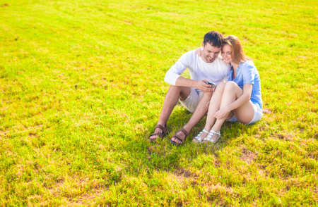 Lifestyle Concept: Caucasian Couple Sitting Embraced Together on the Grass Outdoors with Palmtop Handheld. Listening to Music. Horizontal Image Compositionの写真素材