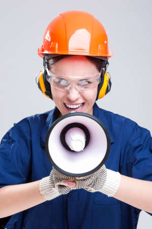 Professions Concepts.Portrait of Expressive Caucasian Female With Loudspeaker Horn Shouting In Hardhat. Equipped with  Coverall. Vertical Image Compositionの写真素材
