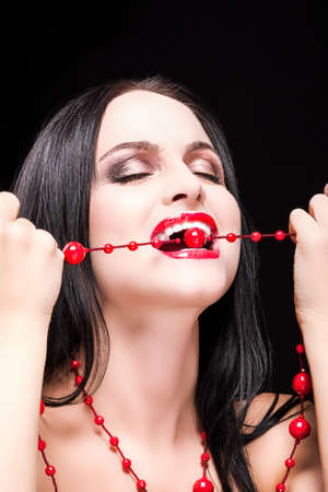 Sensual and Passionate Caucasian Brunette Woman Biting Her Long red Beads. Posing against Black.Vertical Imageの写真素材