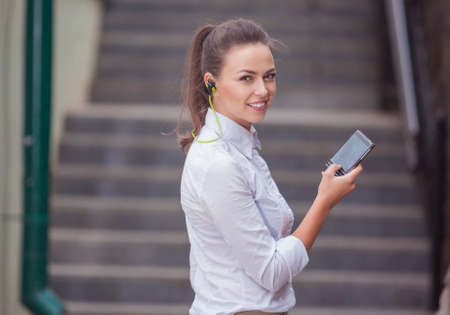 Stylish Modern Caucasian Brunette Woman Posing Outdoors With Smartphone Gadget and Wearing Wireless...