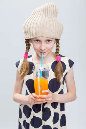Portrait of Funny Caucasian Girl With Pigtails Posing in Warm Hat and Polka Dot Dress with Cup of Orange Juice. Drinking Through Straw. Against White. Vertical image Compositionの写真素材