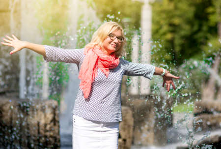 Portrait of Funny Mature Middle Aged Smiling Blond Woman Wearing Spectacles Posing Outdoors in Park. Showing Outstretched hands Against Nature Background.Horizontal Imageの写真素材