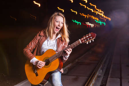 Smiling Caucasian Blond Woman Playing the Guitar Outdoors at Dusk. Mixture of Flash and Halogen...