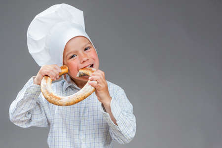Portrait of Happy and Delighted Caucasian Little Boy Eating Fresh Doughnut Bread Roll. Posing In Cooking  Hat Agaunst Gray Background. Horizontal Imageの写真素材