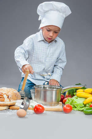 Portrait of Caucasian Boy Working With Kitchen ware In Cooking Hat. Surrounded by Fresh Fruits and Vegetables In Studio. Posing with Smiling Expression. Against Gray.Vertical Image Orientationの写真素材