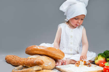 Cooking Ideas and Concepts. Portrait of Little Caucasian Girl in Cook Hat and Lips in Flour Sitting in Front of Fresh Buns and Fruits.Against Gray. Horizontal Imageの写真素材