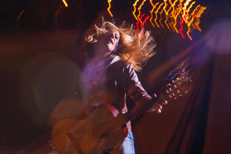 Positive Caucasian Woman In Leather Jacket and Blue Jeans Playing on Rails With Bag Outdoors at Night.Playing the Guitar with Expression. Halogen and Flash Lights are Used. Horizontal Imageの写真素材