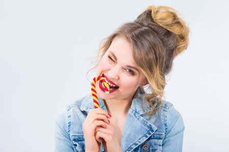 Food Ideas and Concepts. Portrait of Young Surprised Caucasian Blond Girl Eating Big Red-Yellow Candy Cane. Furious Facial Expression. Against White Background. Horizontal Compositionの写真素材
