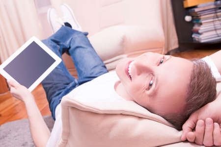 Techology Ideas. Funny Smiling Handsome Man with Tablet Computer Laying Down and Looking Backwards. Legs Lifted on Couch in Front.Focus on Face. Horizontal Imageの写真素材